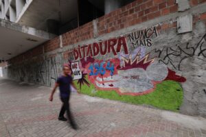 Esta imagem mostra uma parede de tijolos com grafite. O grafite é composto de palavras e números com uma mensagem política e histórica, incluindo as frases DITADURA NUNCA MAIS e NÃO AO GOLPE, acompanhadas do ano 1964 e outros textos como IVAN - JONAS. A parede tem outras pichações e grafites ao longo dela. Na frente da parede, na calçada, há um homem de roupas escuras caminhando, mas ele está desfocado, capturado enquanto está em movimento.
